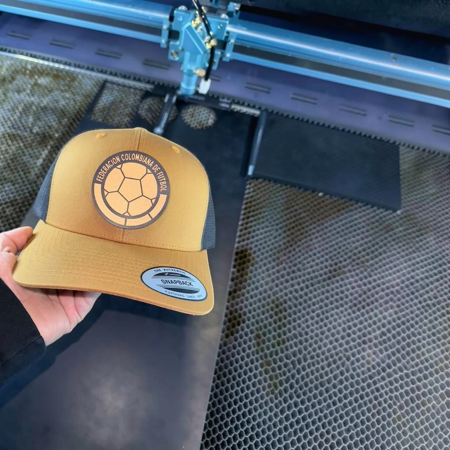 ColChiTex LLc custom engraved patch trucker hat in mustard yellow with a dark grey mesh back and a circular patch featuring a soccer ball and text.